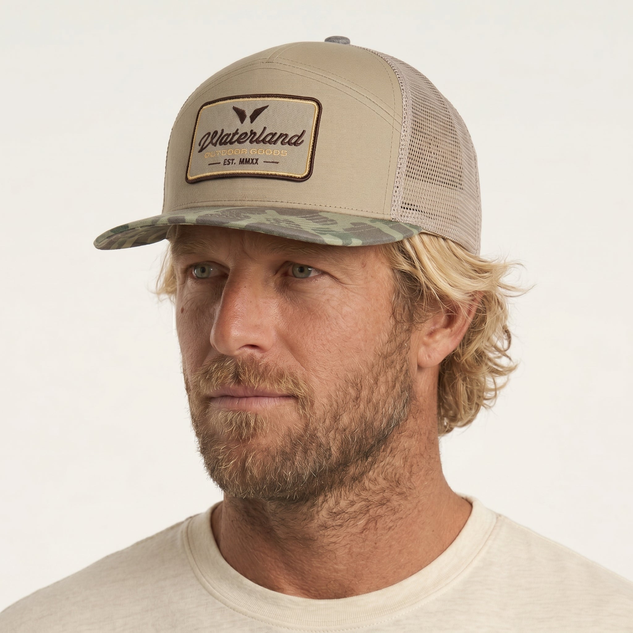 Man wearing a beige and camouflage trucker hat with a visible brand logo on a white background